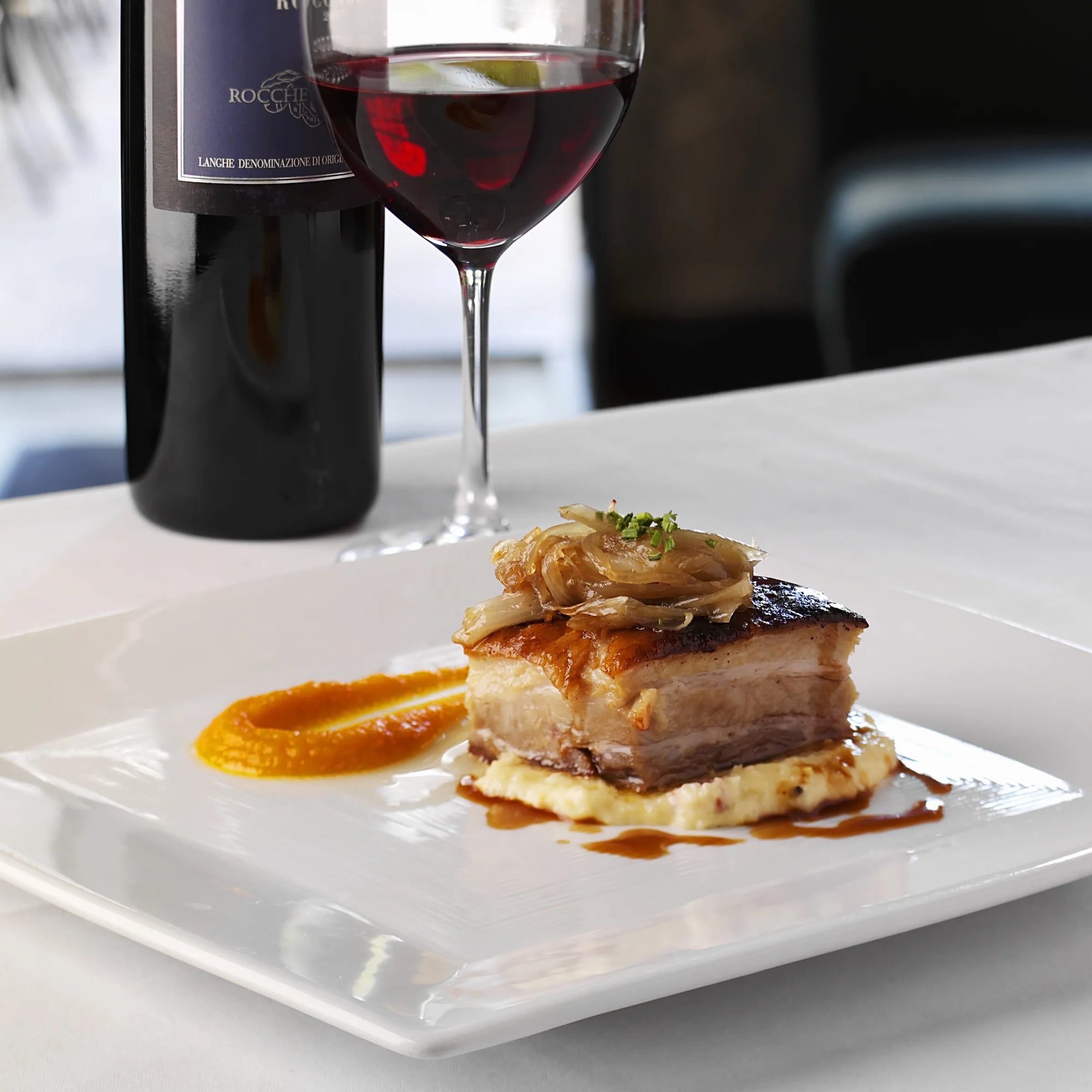 Meat on a white platter with sauce and a glass of red wine on table