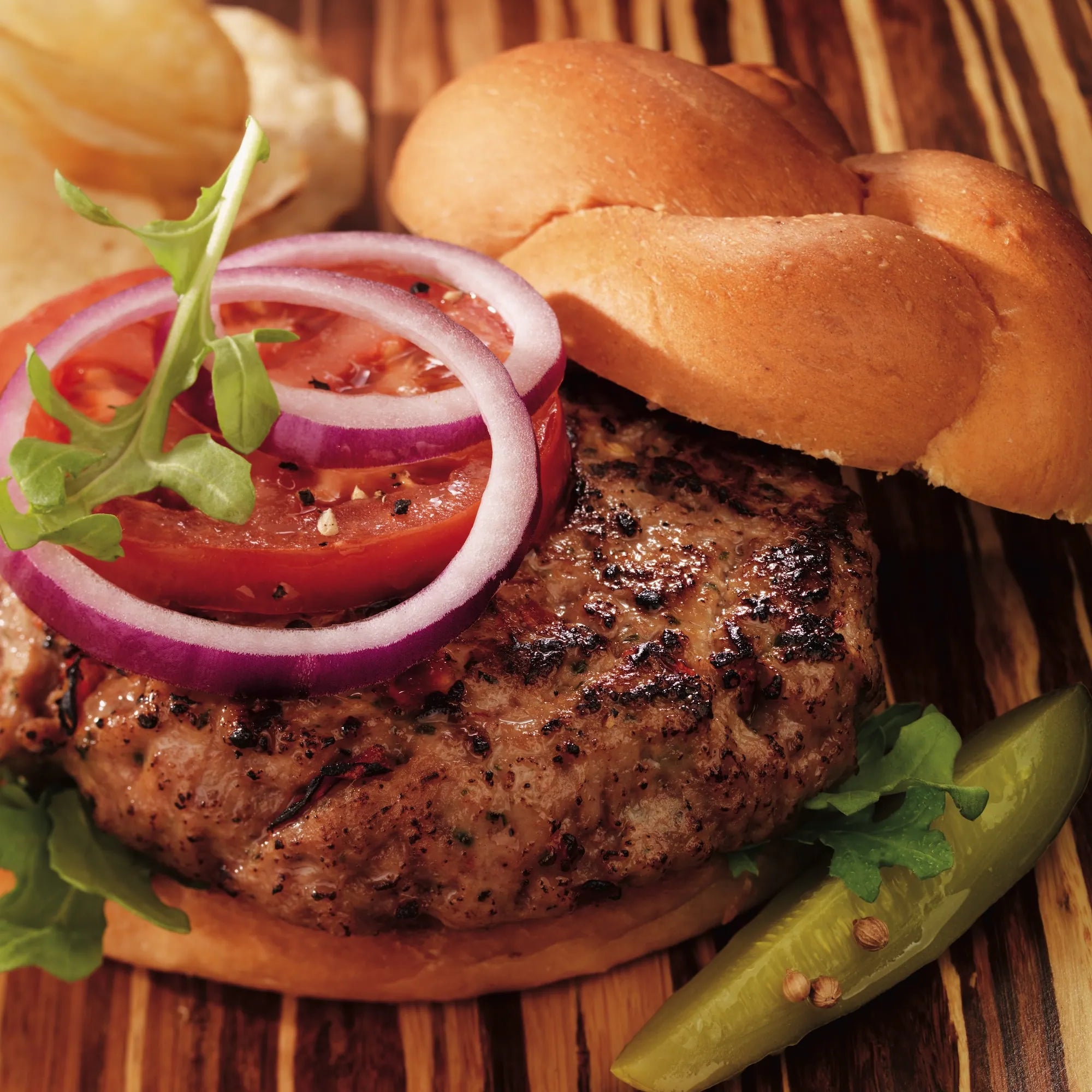 Turkey Burger on a bun with arugula, tomato, and red onion, alongside a pickle spear and potato chips, highlighting its grilled, seasoned appeal.