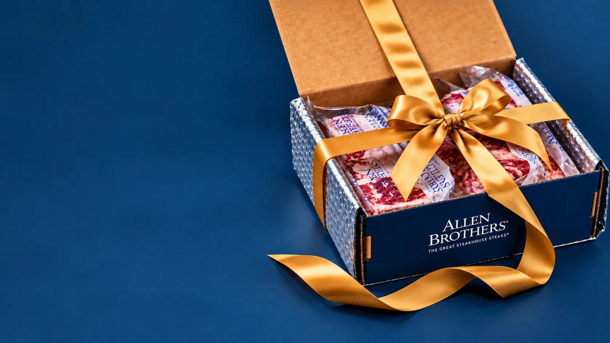 An open Allen Brothers gift box tied with a gold ribbon, showing packaged gourmet meats inside, set against a dark blue background