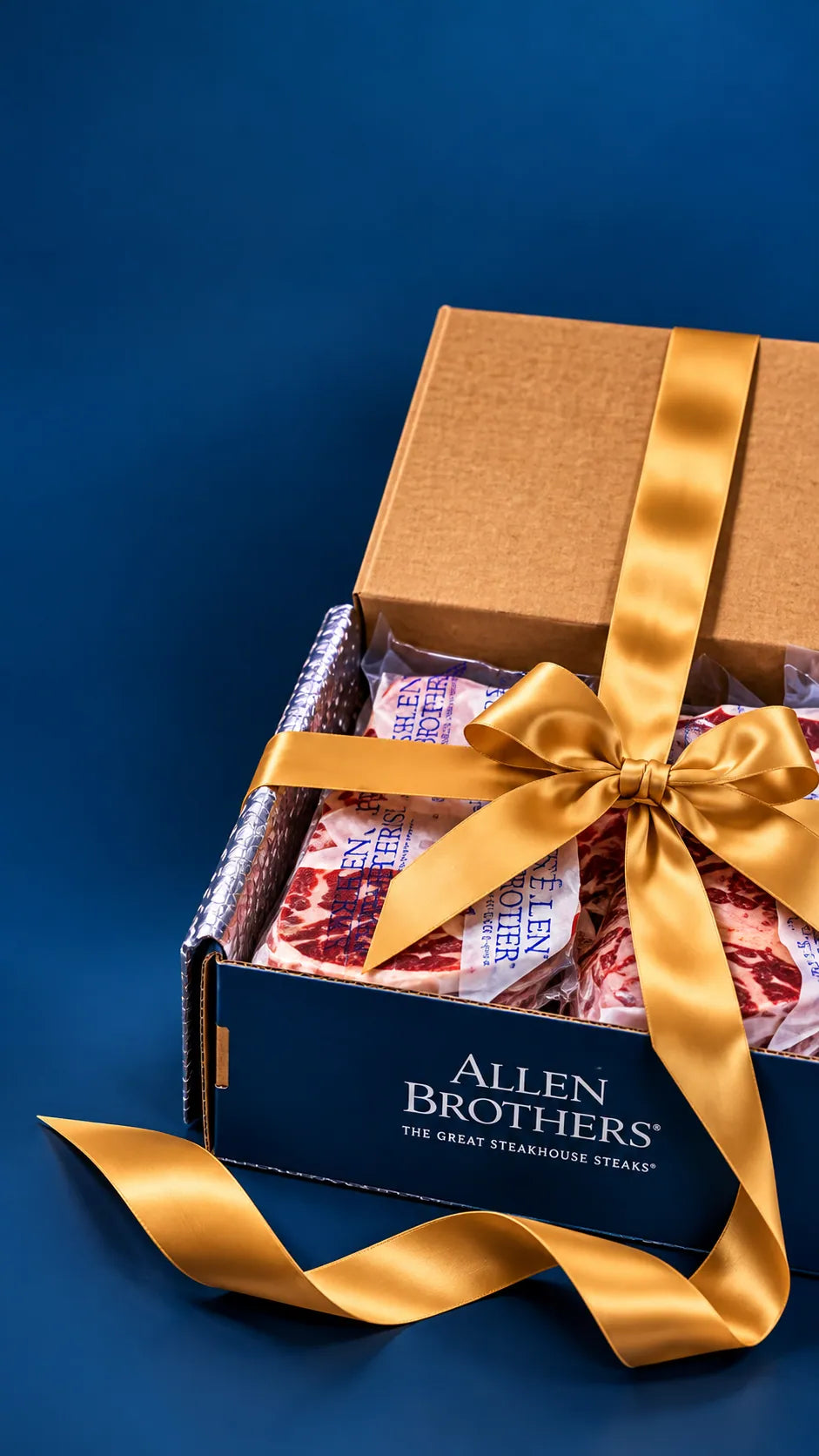 An open Allen Brothers gift box tied with a gold ribbon, showing packaged gourmet meats inside, set against a dark blue background