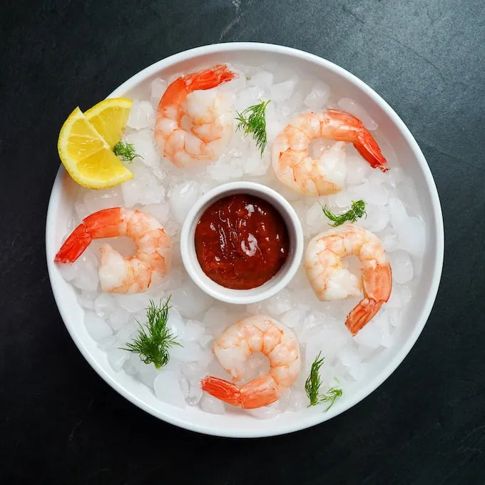 Jumbo Cooked Shrimp on ice with tails on, featuring six shrimp, a central cocktail sauce ramekin, lemon wedge, and dill garnish, perfect for appetizers.