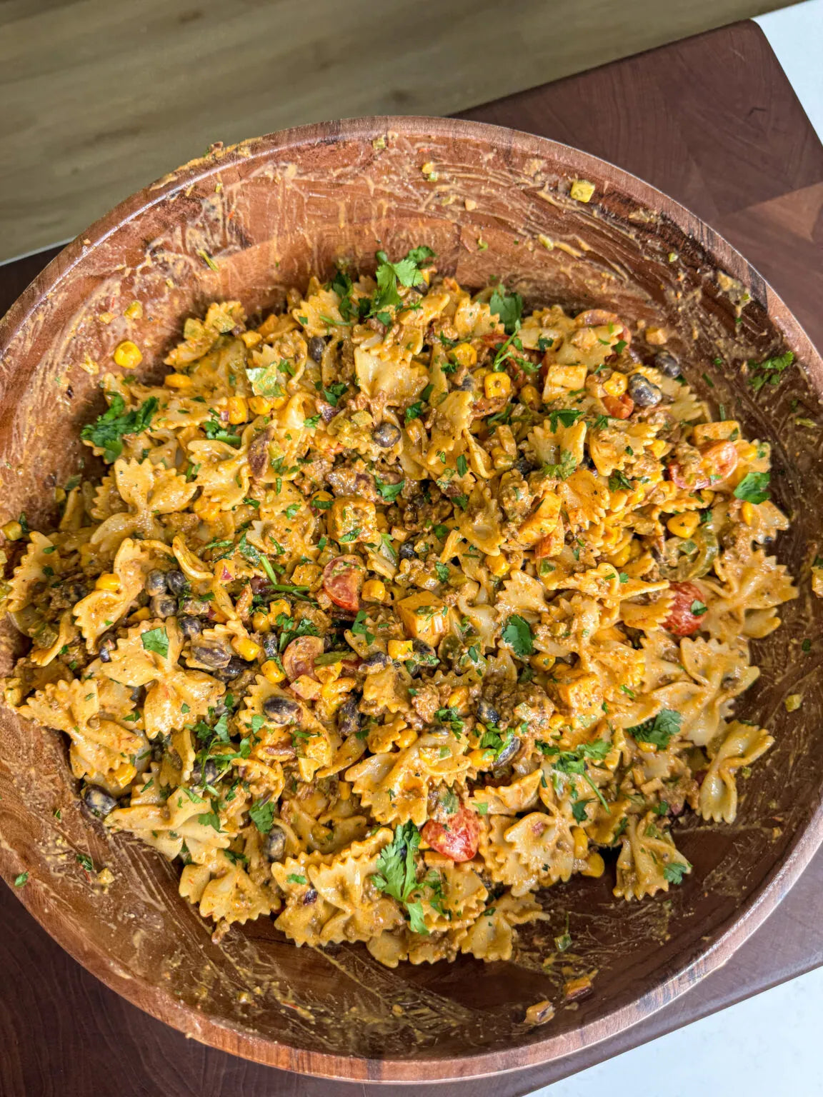 A top-down view of a creamy pasta salad in a large wooden bowl, featuring bowtie pasta mixed with corn, black beans, diced tomatoes, and fresh cilantro.