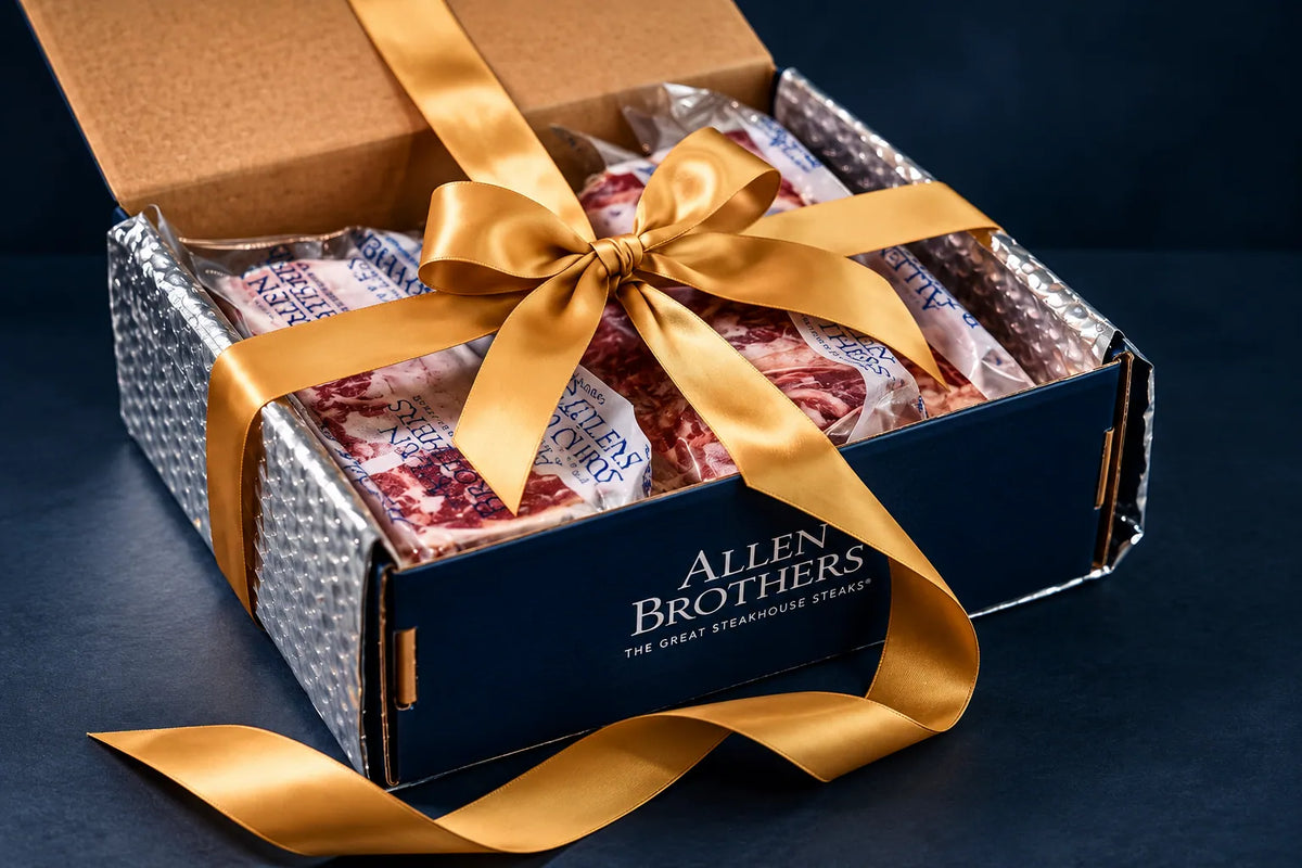 An open Allen Brothers gift box containing packaged steaks, wrapped in a shiny gold ribbon and tied in a bow, set against a dark navy background.