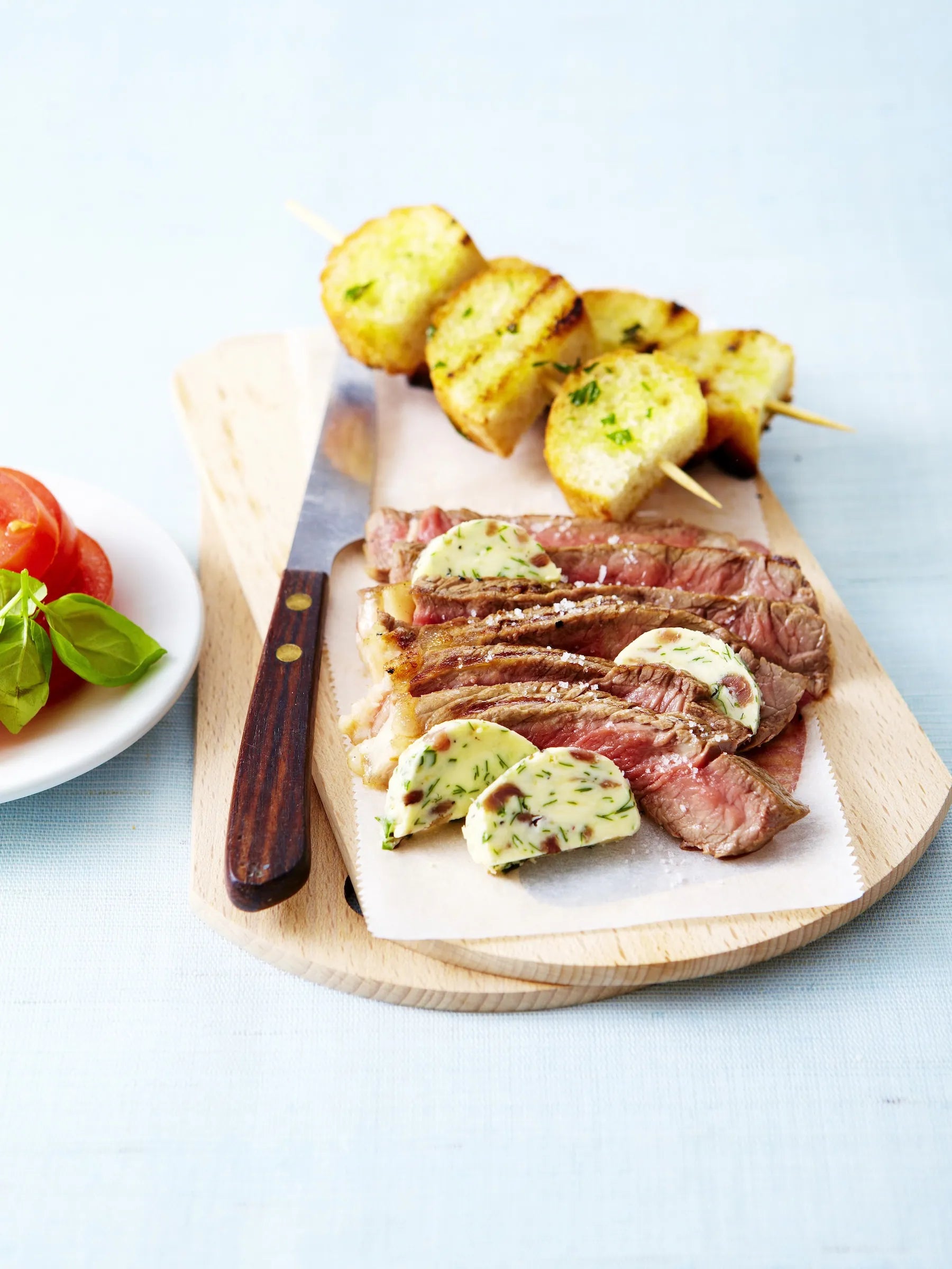 Steak with garlic butter and potato skewers on a wooden board with a knife.
