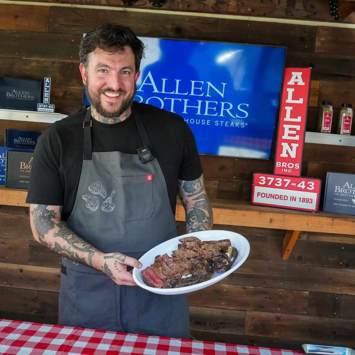 A smiling man with tattoos wearing a gray Allen Brothers House Steaks apron stands behind a counter holding a white plate of cooked meat. The background features a wooden wall, a blue Allen Brothers sign, and a red and white checkered tablecloth in the foreground.