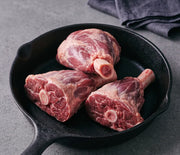 Domestic All Natural Volcano Lamb Shank in a skillet, featuring three raw shanks with exposed frenched bones, ideal for slow-braising or osso buco.