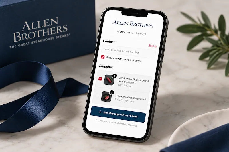A smartphone displaying the Allen Brothers restaurant app on a light marble surface, with a navy Allen Brothers gift box and ribbon, a small green plant, and part of a white plate in the background.
