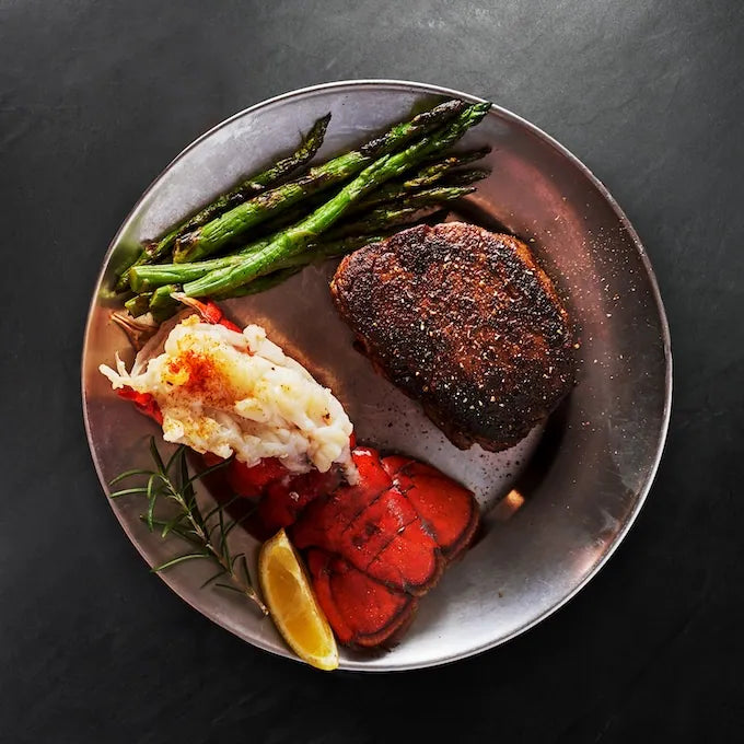 Surf & Turf XL Combo featuring pepper-crusted steak, butterflied lobster tail, grilled asparagus, with a lemon wedge and rosemary garnish.